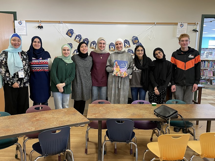 Group of volunteers posing in a library setting, smiling and holding a children's book about Ramadan, with a decorative banner in the background.
